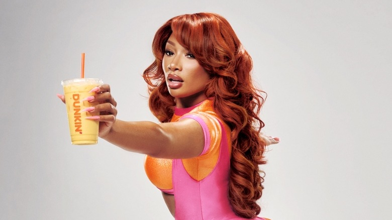 Megan Thee Stallion portant un équipement d'entraînement rose et orange faisant une fente et tenant un rafraîchisseur de protéines Megan's Mango