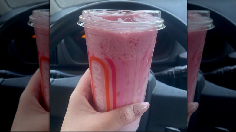 Main de femme tenant un Ultimate Pink Daydream Refresher dans une tasse Dunkin en plastique devant un volant
