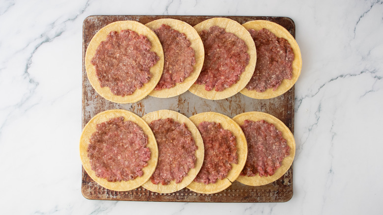 Une plaque à biscuits remplie de tortillas de maïs avec des galettes de hamburger écrasées dessus