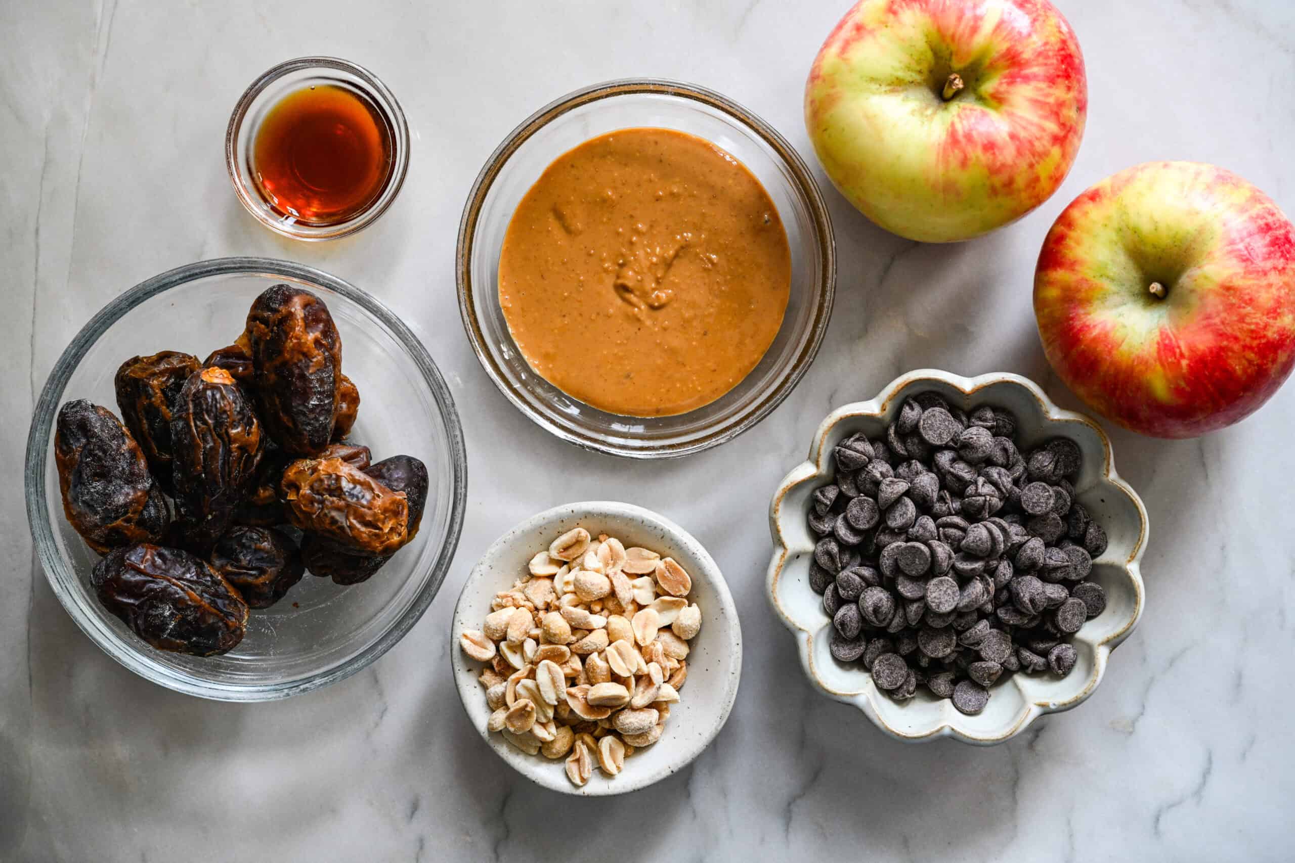 Vue aérienne des dattes, du beurre de cacahuète, de la vanille, des cacahuètes, des pépites de chocolat et de deux pommes sur un comptoir en marbre.