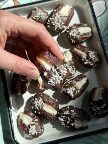 Vue rapprochée d'une main soulevant une datte enrobée de chocolat farcie de noix de coco dans un plateau.