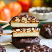 Vue de face de deux tranches de pomme enrobées de chocolat coupées en deux, empilées pour montrer des couches de pomme, de dattes et d'arachides, avec des pommes derrière.