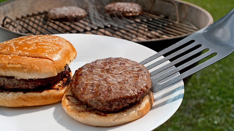 Spatule plaçant un hamburger sur un petit pain sur une assiette blanche avec un gril au charbon de bois en arrière-plan