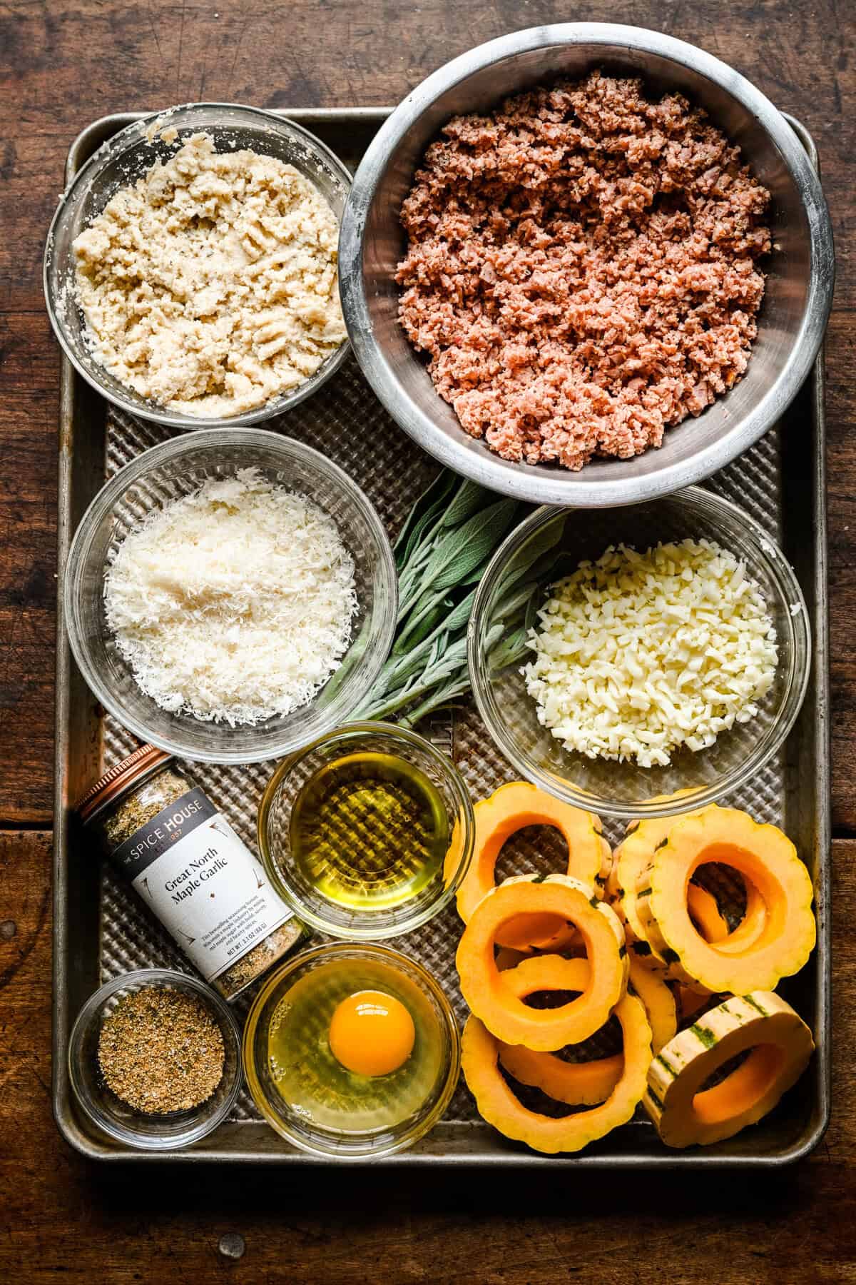 Vue aérienne des ingrédients sur une plaque à pâtisserie : saucisse hachée, chapelure, parmesan, mozzarella, sauge fraîche, huile d'olive, un œuf, assaisonnement à l'ail Great North Maple et rondelles de courge delicata.