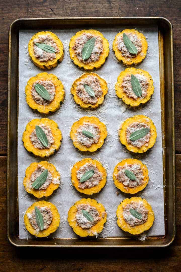 Vue aérienne des rondelles de courge sur une plaque remplie de saucisses, saupoudrée de parmesan et garnie de feuilles de sauge avant la cuisson.