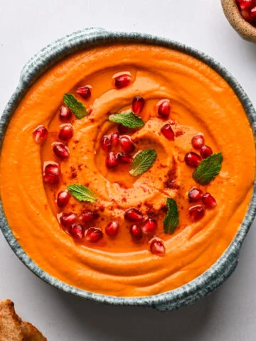 Vue aérienne de la trempette feta aux carottes et aux poivrons rouges rôtis dans un bol bleu, garnie de graines de grenade et de menthe, avec du pain pita et de petits bols de graines de grenade et de menthe sur le côté.