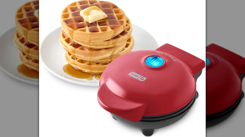Petit gaufrier Dash rouge avec une lumière bleue allumée sur le dessus à côté d'une pile de gaufres sur une assiette blanche avec du sirop et du beurre
