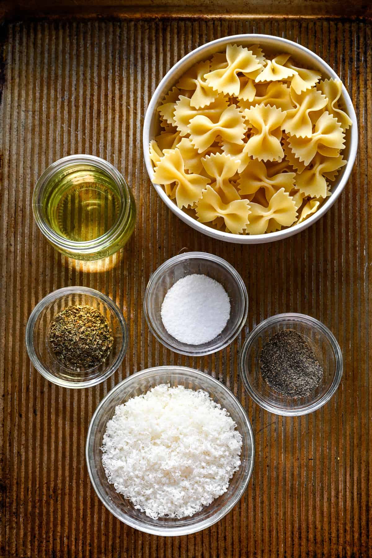 Vue aérienne des ingrédients des pâtes feuilletées sur une plaque à pâtisserie : pâtes à nœud papillon, huile, herbes séchées, sel, poivre et parmesan râpé.