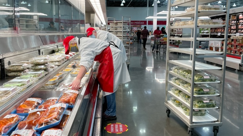 Deux employés de Costco réapprovisionnent des contenants de charcuterie dans un réfrigérateur à portée de main à l'intérieur d'un magasin-entrepôt avec un chariot derrière eux.