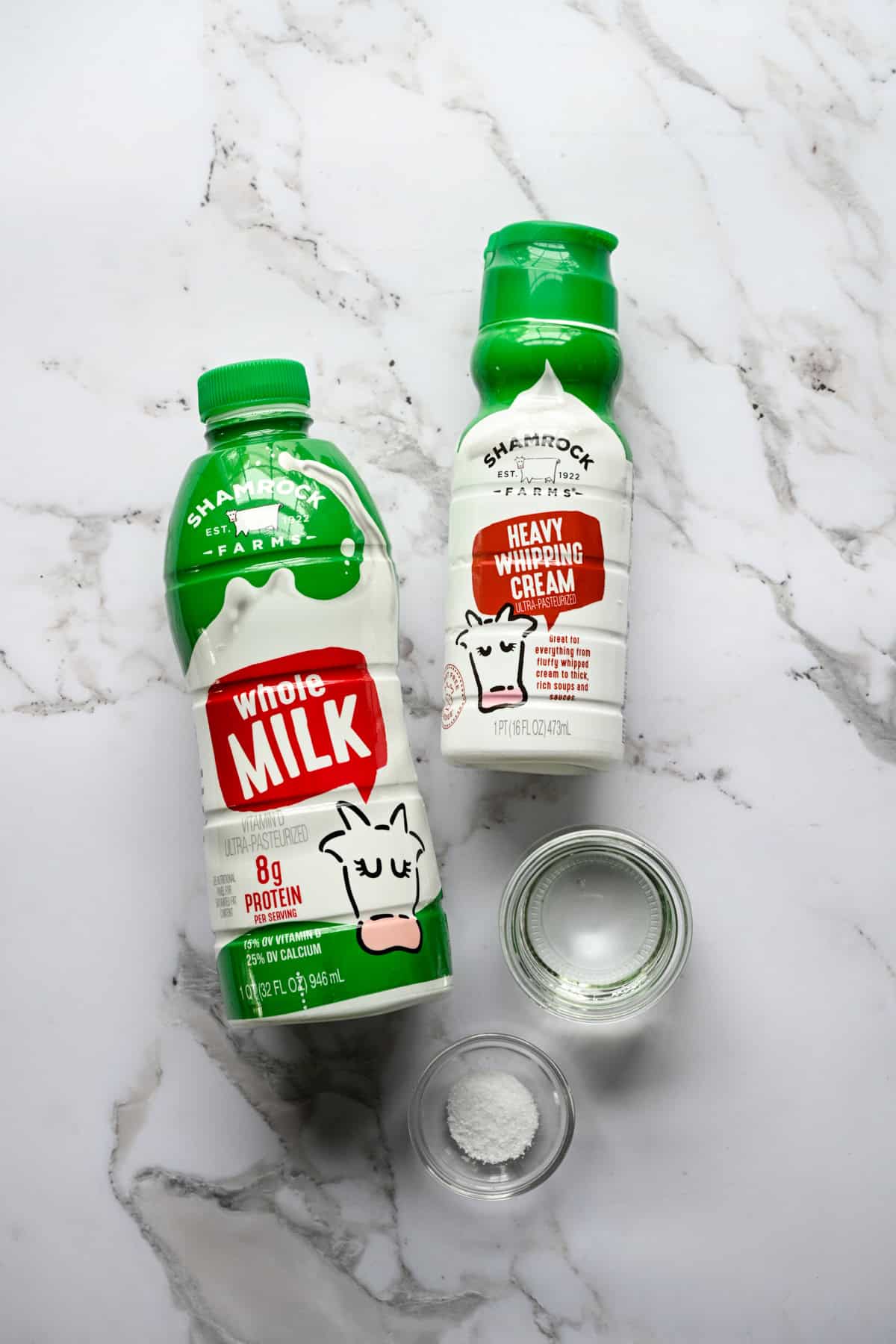 Vue aérienne des ingrédients du fromage cottage fait maison : une bouteille de lait entier Shamrock Farms, de la crème fouettée épaisse Shamrock Farms et de petits bols en verre de vinaigre blanc et de sel sur du marbre