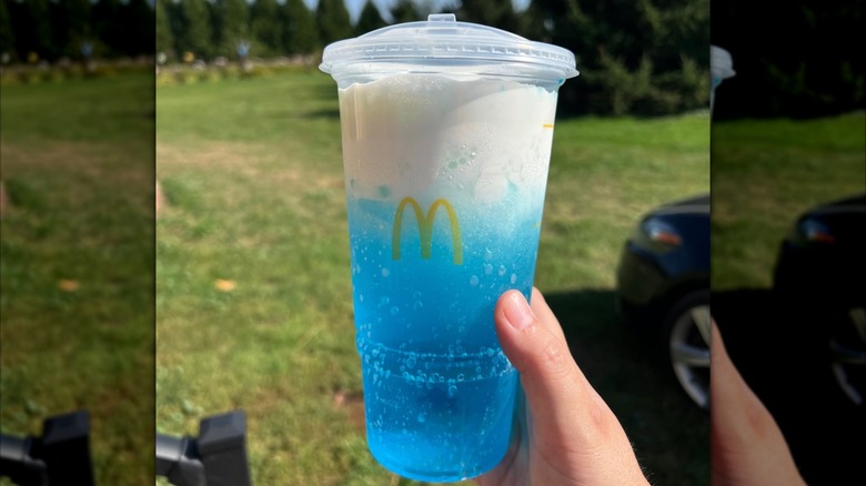 La main d'une personne tenant un McDonald's Sprite Berry Blast bleu et blanc recouvert de mousse sur un fond d'arbres et d'herbe